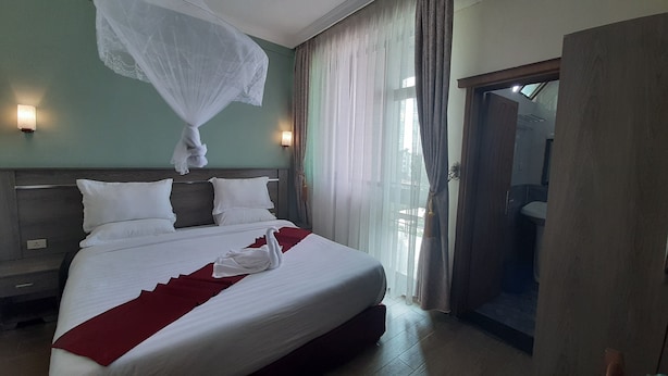 A hotel room with a large bed, a nightstand, a wall-mounted lamp, and a view of the outside through a window with curtains.