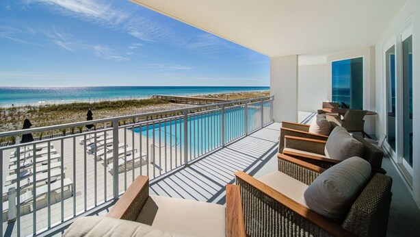 A balcony with a view of the ocean, a pool, and outdoor seating.