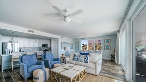A modern living room with a sofa, chairs, a coffee table, and a dining area with a table and chairs. There is a kitchen in the background with cabinets and appliances.