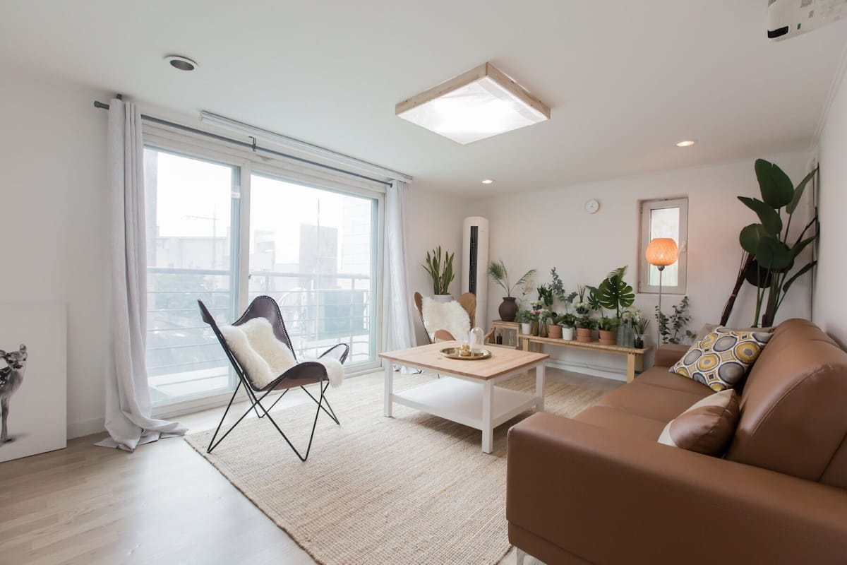 A modern living room with a brown leather sofa, a white coffee table, and a black folding chair. There is a large window with sheer curtains, a potted plant, and a floor lamp.