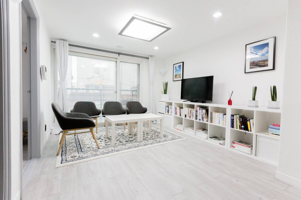 A modern living room with a white color scheme, a flat-screen TV, a bookshelf, and a dining area with chairs and a table.