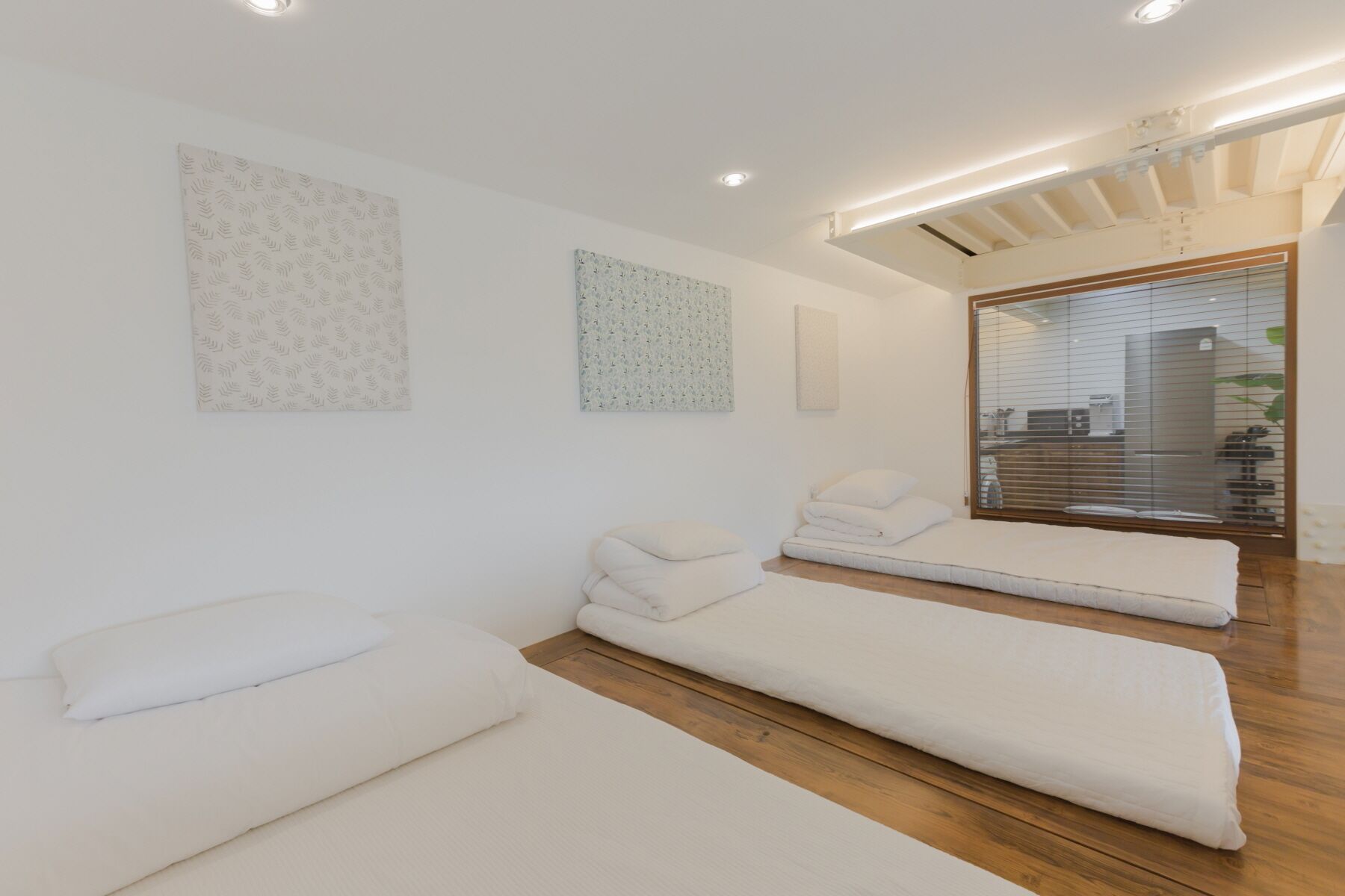 A minimalist room with three white beds, wooden flooring, and a view of a kitchen through a glass door.