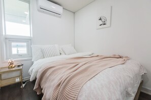 A bedroom with a bed, white bedding, a pink knitted blanket, a small bedside table with a lamp, and a wall-mounted air conditioner. - Sio R-Suite (Seoul)