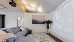 Ein modernes Wohnzimmer mit einem grauen Sofa, einem gemusterten Teppich, einer Projektionsleinwand und einem gerahmten Landschaftsbild.