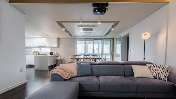 Ein modernes Wohnzimmer mit einem grauen Ecksofa, einem kleinen Hocker und einem gemusterten Teppich.