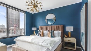 Ein modernes Schlafzimmer mit einem großen Bett, zwei Nachttischen, einem runden Spiegel und Blick auf eine Stadtlandschaft durch ein großes Fenster.