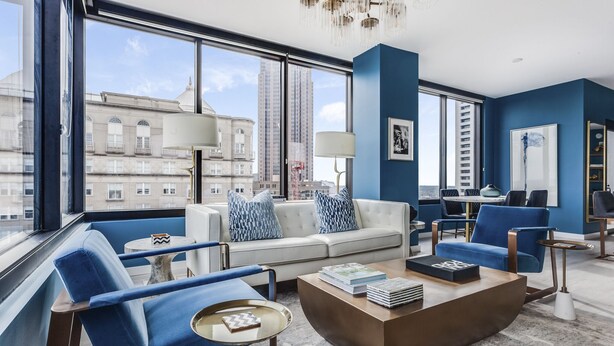 Ein modernes Wohnzimmer mit einer Couch, Sesseln und einem Couchtisch. Große Fenster bieten Blick auf die Stadt.