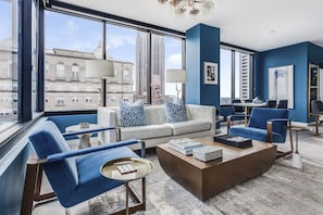 A modern living room with a sofa, armchairs, and a coffee table. Large windows offer a city view.