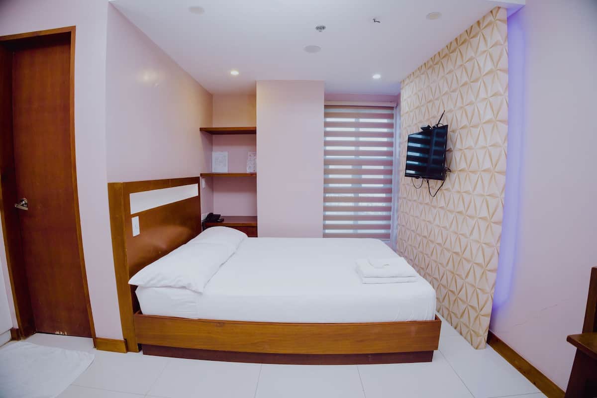 A neatly made bed in a room with a wooden headboard, a wall-mounted TV, and a patterned wall.