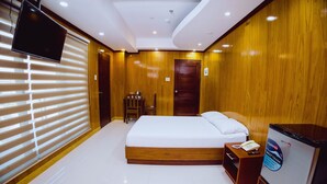 A hotel room with a large bed, wooden paneling, a flat-screen TV, a bedside table with a phone, and a wooden door. - Coastal Hotel Iloilo (Leganes)