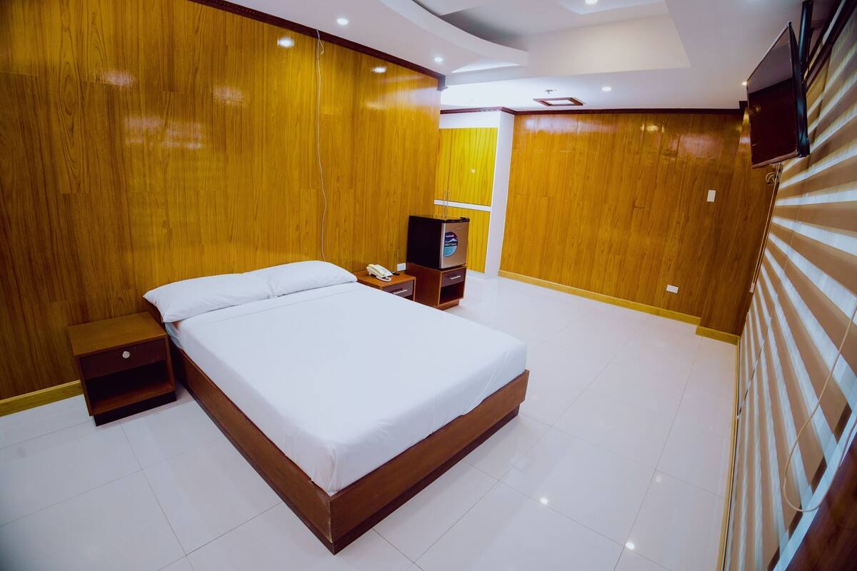 A hotel room with a large bed, wooden panel walls, a small bedside table with a phone, and a television on a stand.