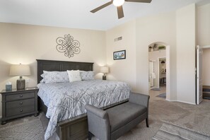 A bedroom with a bed, two bedside tables, a bench, a ceiling fan, and a door leading to another room.