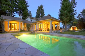 Una casa de dos pisos con piscina, rodeada de árboles y un patio de piedra.