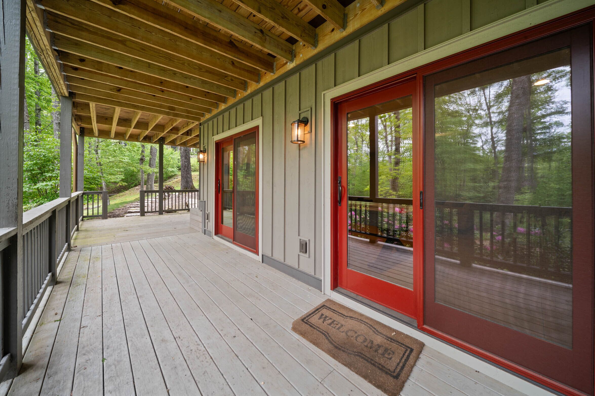 A covered porch with a wooden deck, red-framed glass doors, and a welcome mat.