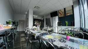 A modern dining area with multiple tables and chairs, a kitchen area in the background, and a view of the outside through large windows. - Hôtel du Tramway (Laon)
