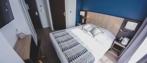 Habitación de hotel con una cama grande, una mesita de noche con lámpara, cabecero de madera y vistas al pasillo.