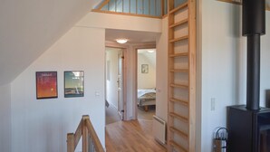 Una escalera lleva a un dormitorio en entrepiso con una escalera de madera, un calentador negro y dos cuadros en la pared.
