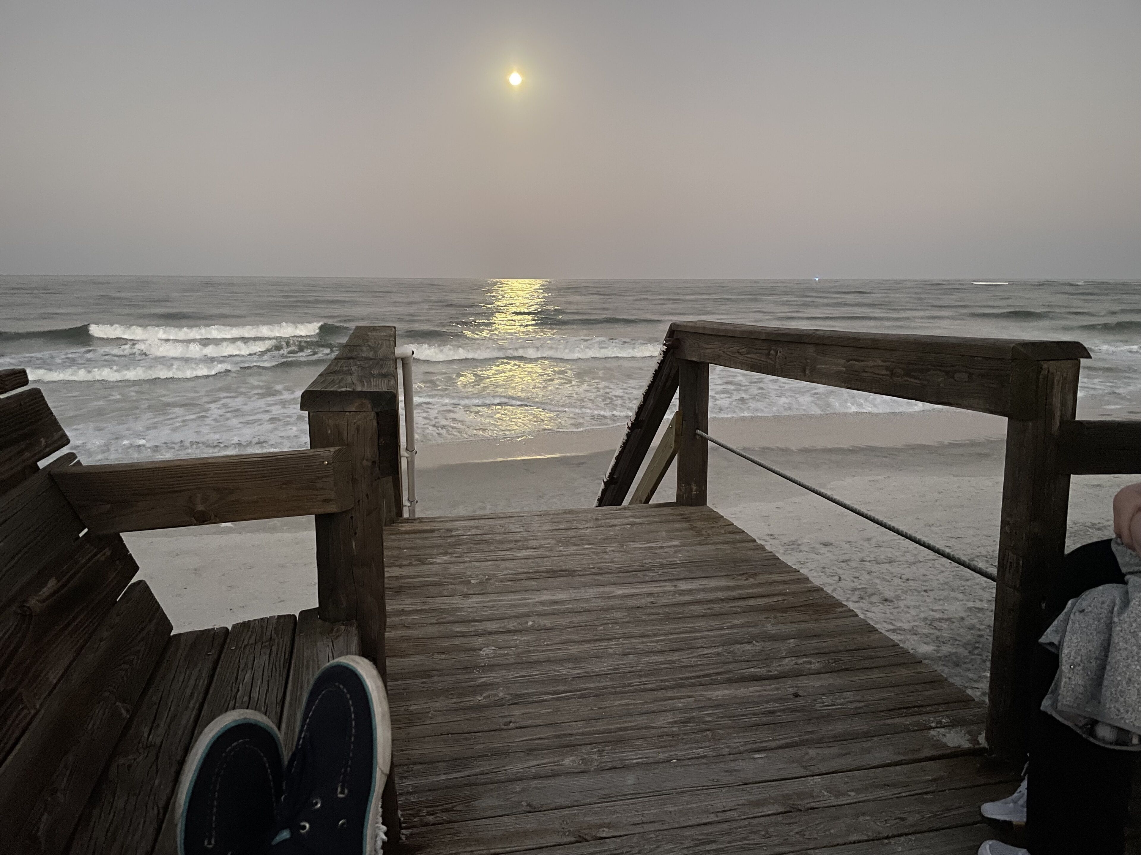 A wooden boardwalk leading to a beach with waves and a sun in the background.