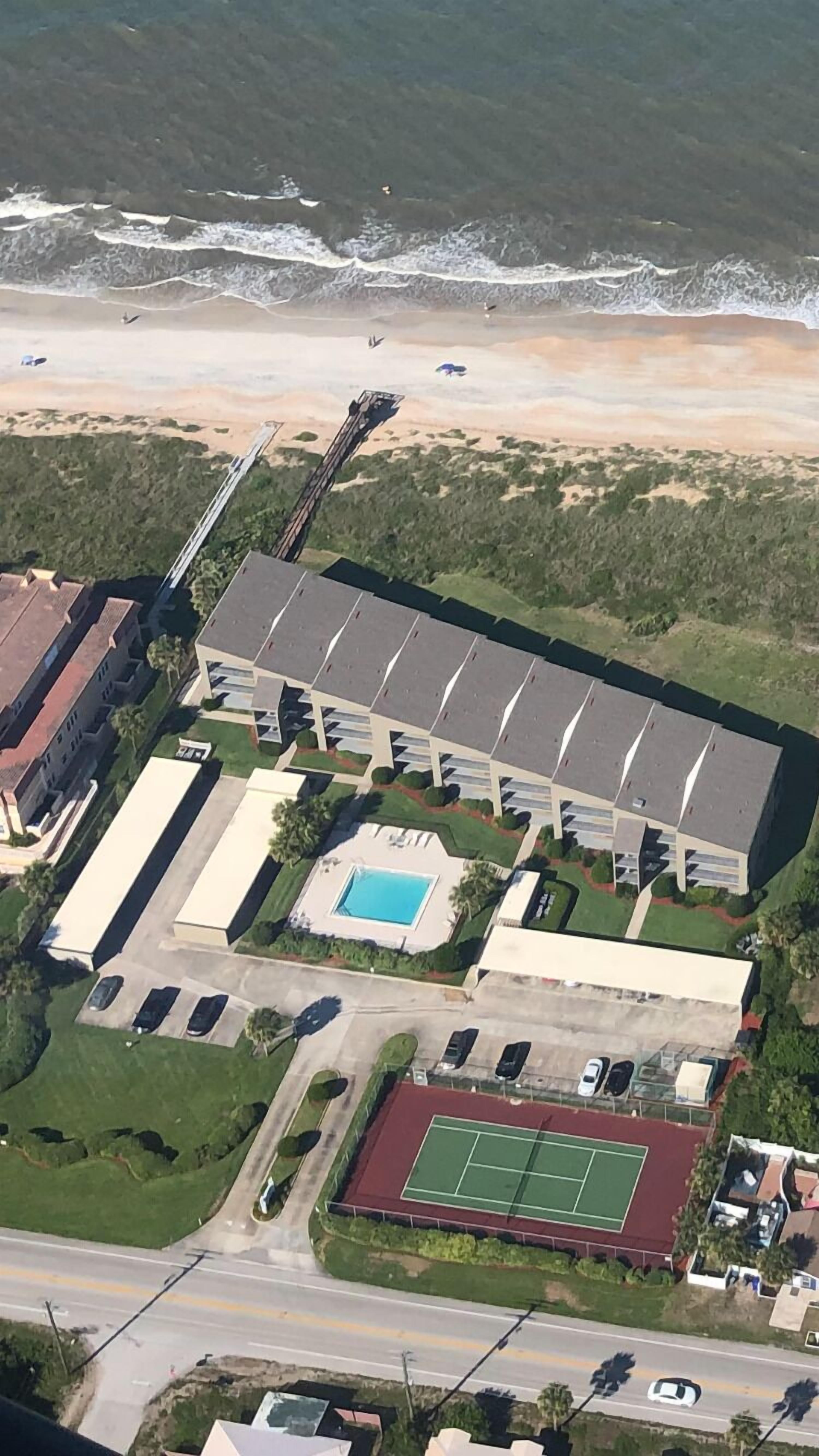 An aerial view of a large building with a swimming pool, tennis court, and parking area.