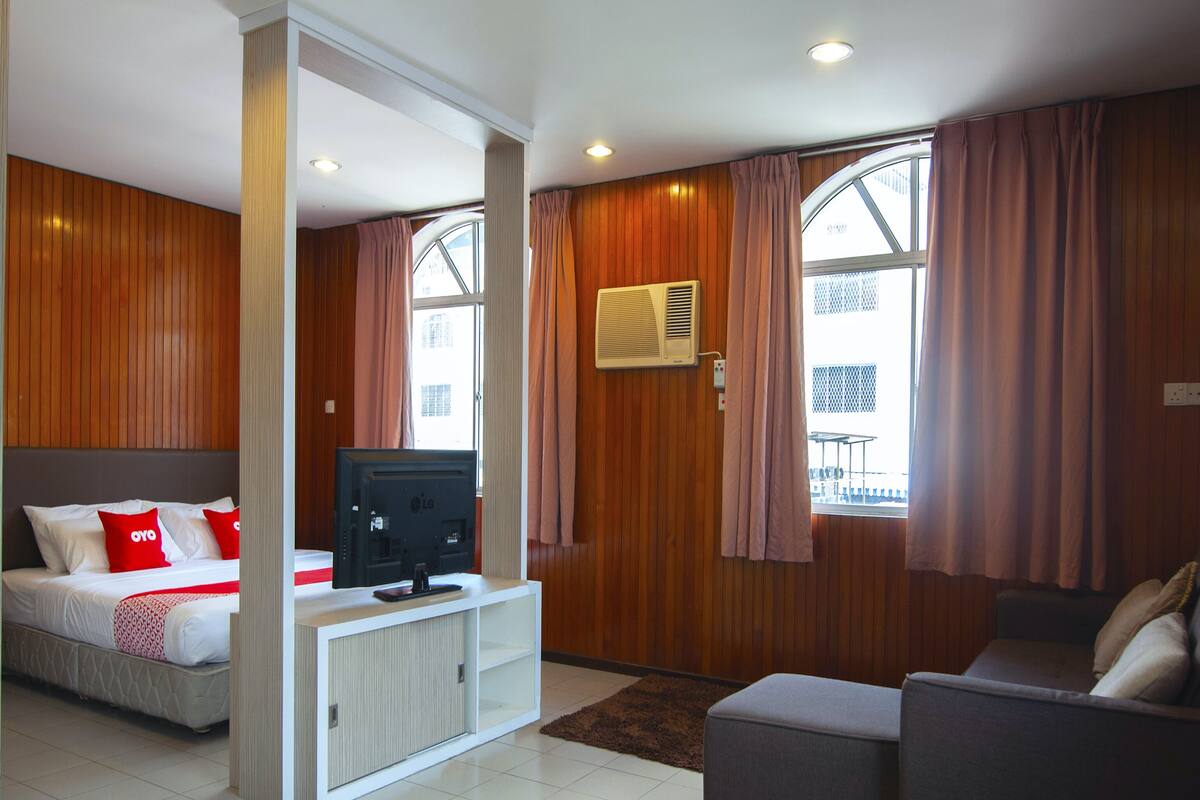 A hotel room with a bed, a TV, a sofa, and large windows with curtains.