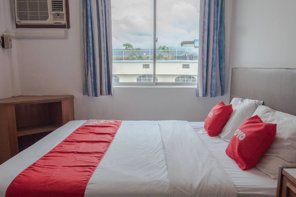 A hotel room with a bed, a nightstand, a window with curtains, and an air conditioning unit on the wall.