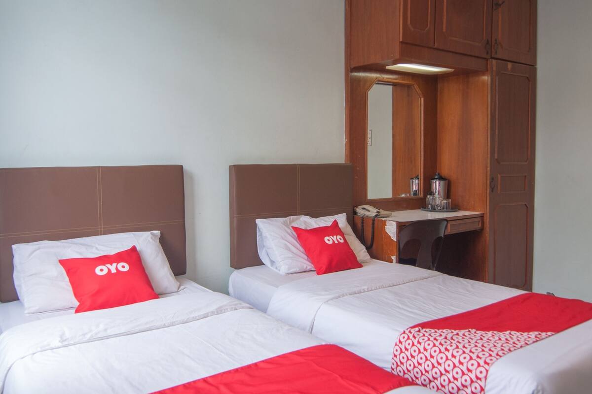 A hotel room with two beds, each with white bedding and red pillows. There is a wooden wardrobe and a small desk with a mirror above it.