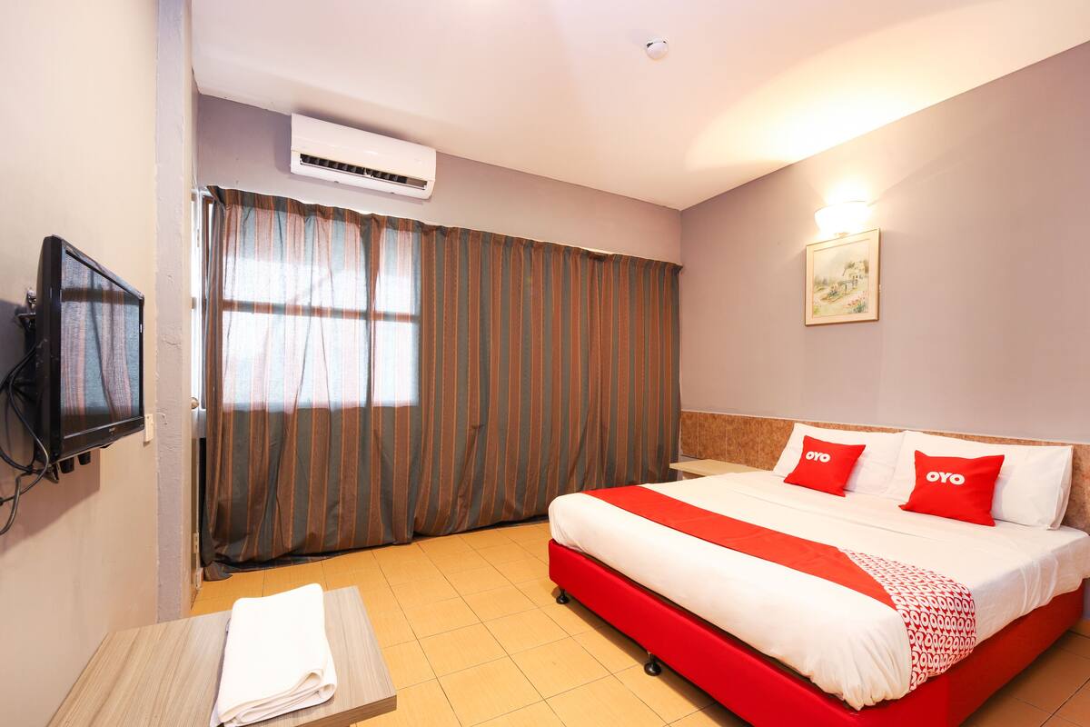 A hotel room with a bed, a TV, an air conditioner, a window with curtains, and a painting on the wall.