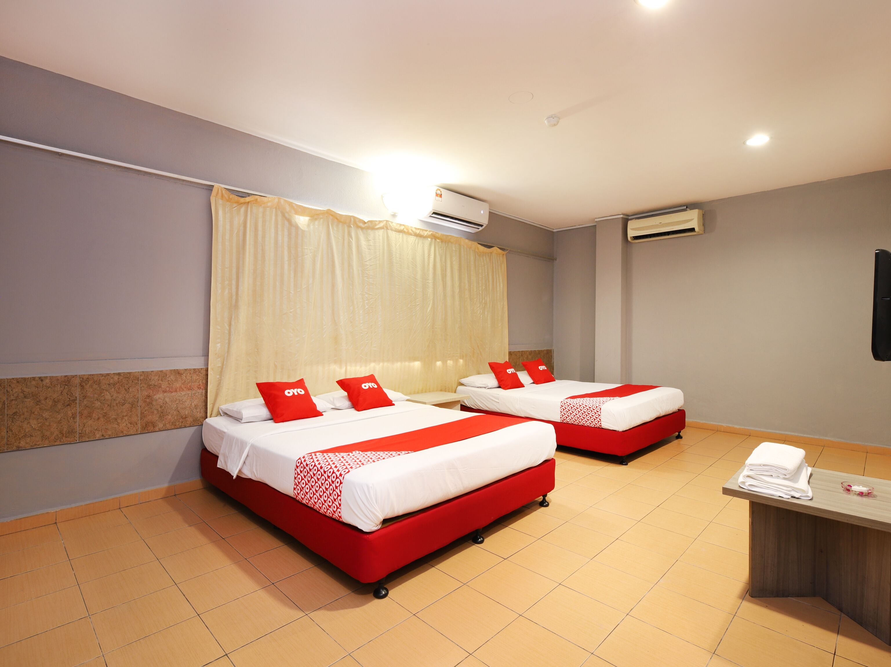 A hotel room with two beds, each with red and white bedding, a wooden desk, and an air conditioning unit on the wall.