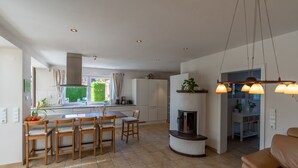 Cocina moderna con una isla central, armarios blancos y estufa integrada. Cuenta con un comedor con sillas de madera y una mesa, además de una sala de estar con sofá y chimenea.