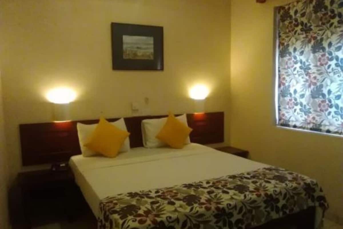 A hotel room with a bed, two bedside lamps, a window with floral curtains, and a framed picture on the wall.