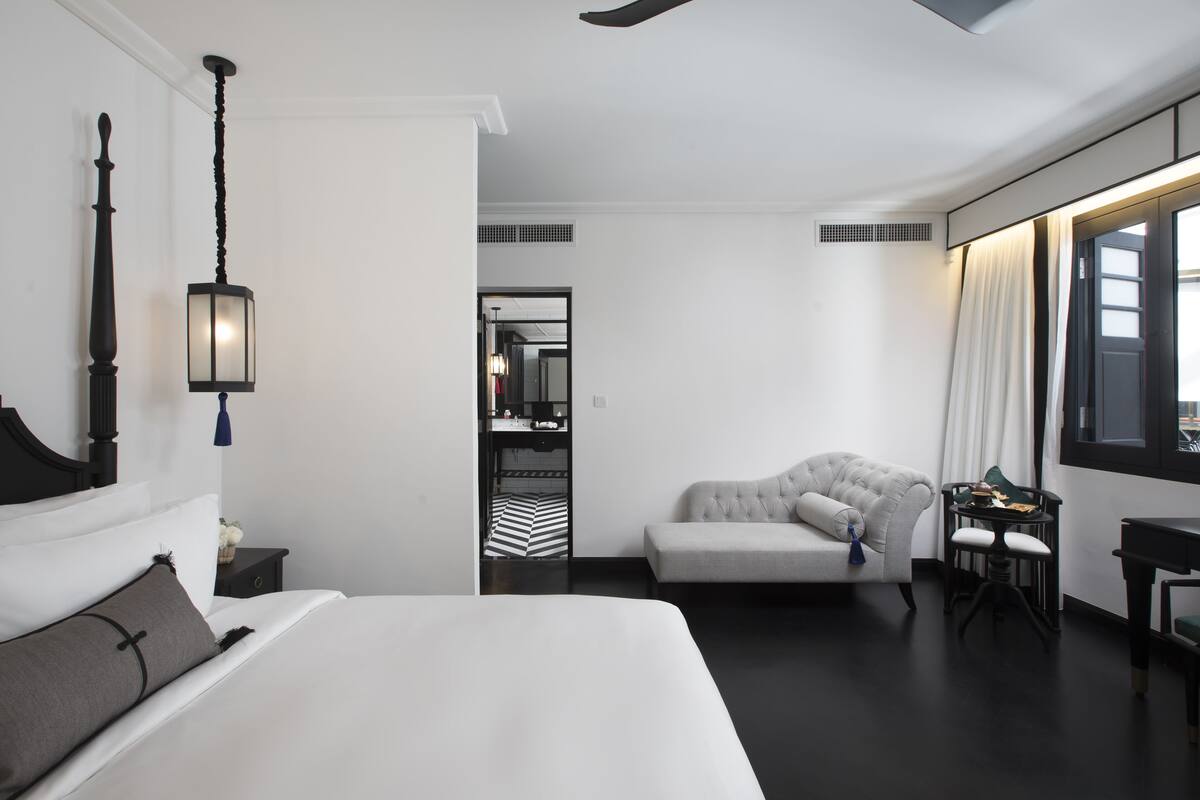 A modern hotel room with a large bed, a seating area with a sofa and armchairs, a small table, and a view of the city through the window.