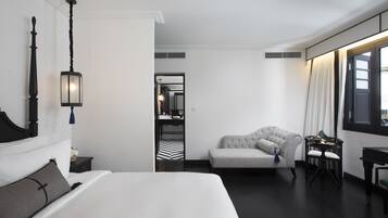 A modern hotel room with a large bed, a seating area with a sofa and armchairs, a small table, and a view of the city through the window.