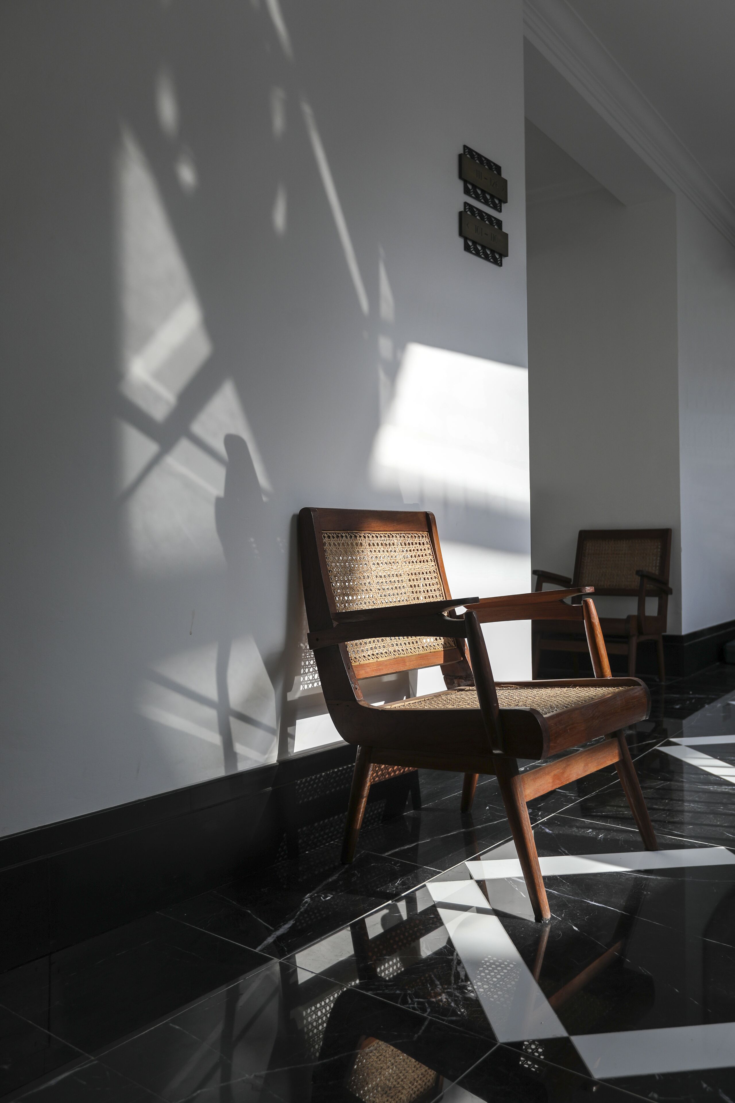 A wooden bench with a woven backrest against a wall, a tiled floor, and a shadow cast by a window.