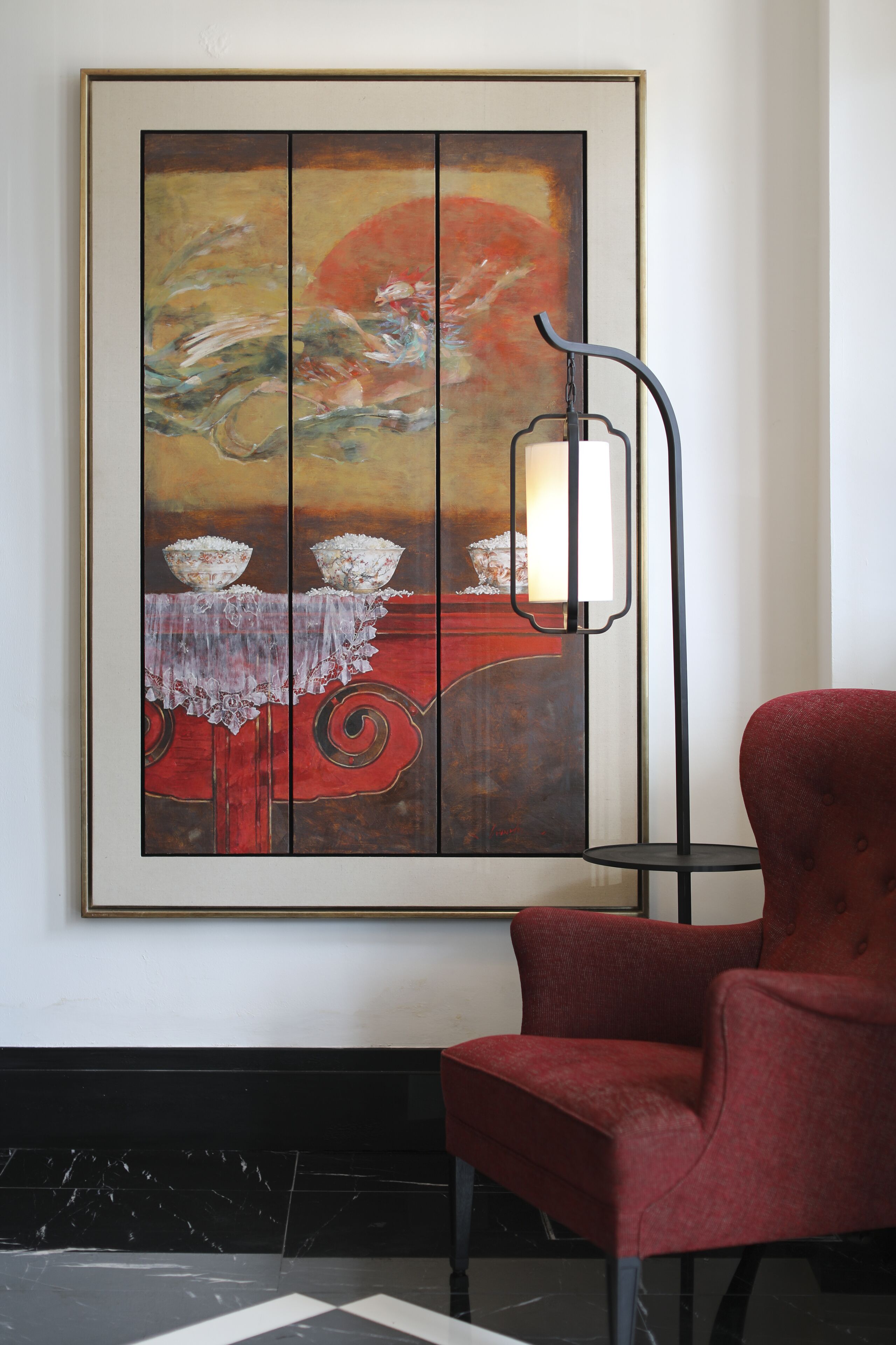 A framed abstract painting on a wall, a red chair, and a floor lamp.
