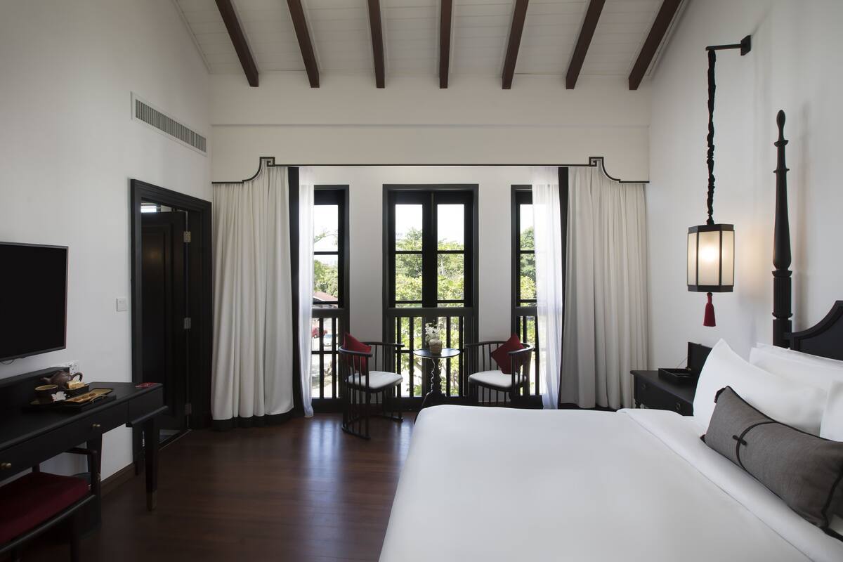 A hotel room with a large bed, a desk with a TV, two chairs, a small table, and a balcony with curtains.