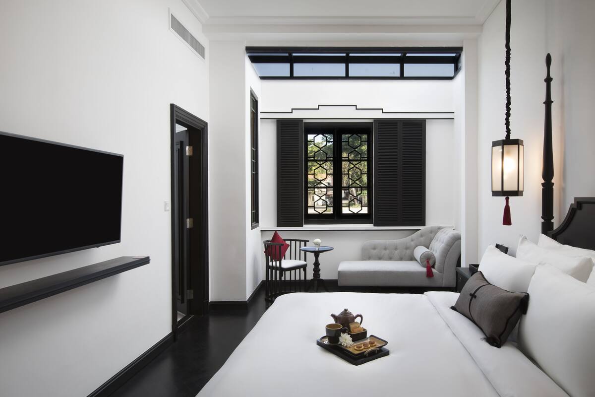A modern hotel room with a large bed, a TV mounted on the wall, a small table with a tray of tea and pastries, a sofa, and a window with black shutters.
