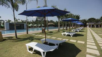 A poolside area with white lounge chairs and blue umbrellas, surrounded by palm trees and a clear sky.