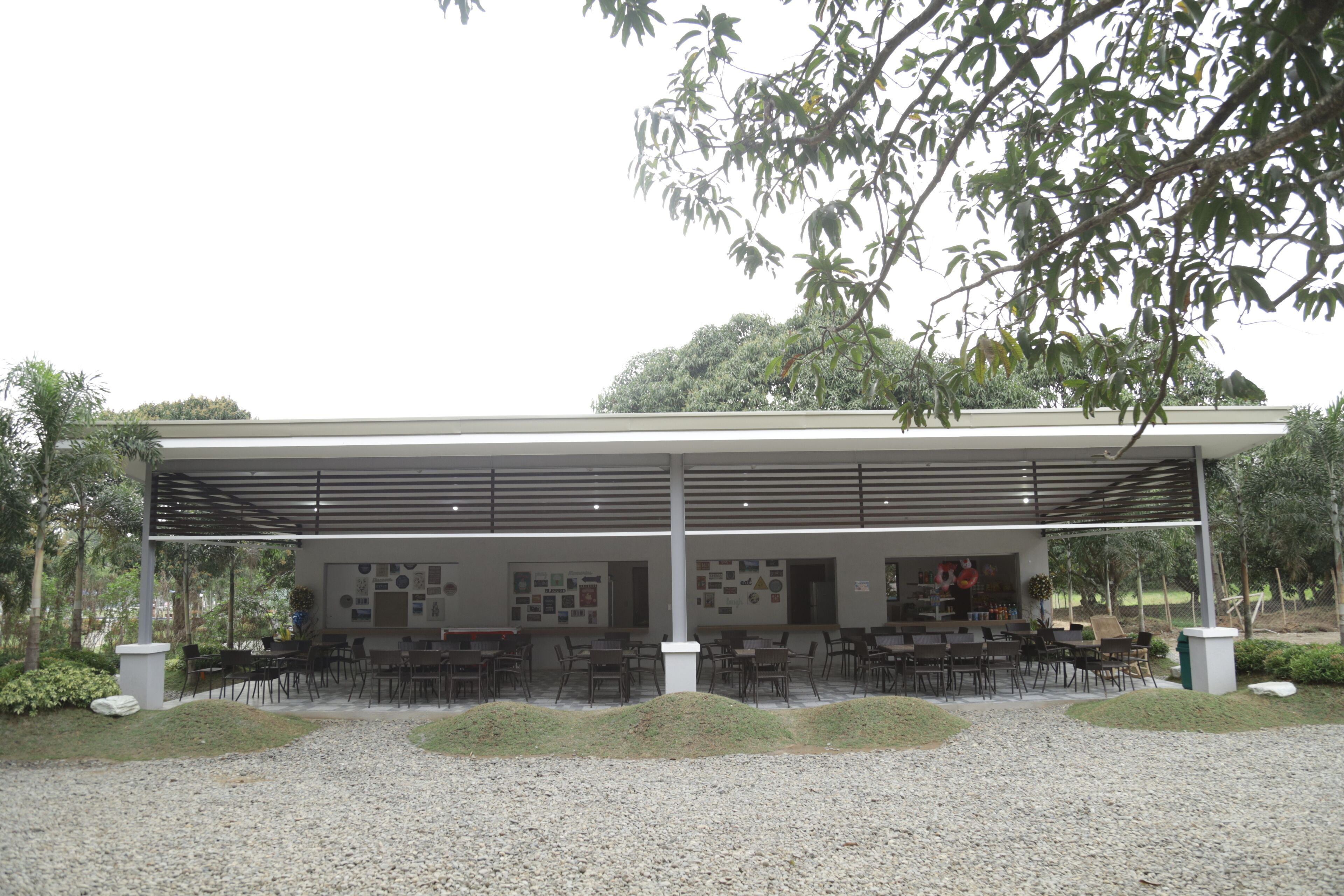 A covered outdoor seating area with multiple tables and chairs, surrounded by greenery and a gravel path.