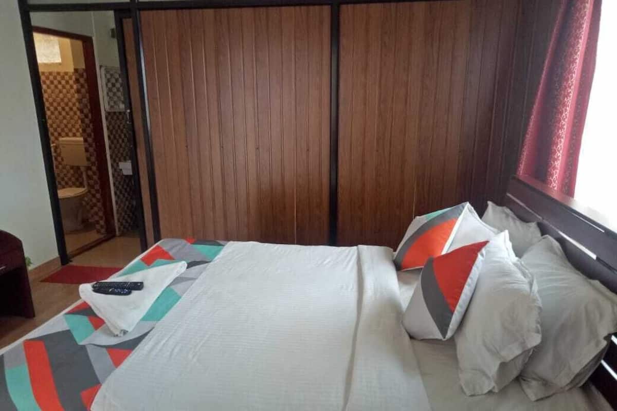A neatly made bed with a white comforter and pillows, a remote control on the bed, and a wooden wardrobe in the background.