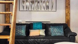 A loft-style room with a wooden ceiling, a black leather sofa with patterned cushions, a white table, and a metal wall art piece. - RoHouse delle Conserve (Arezzo)