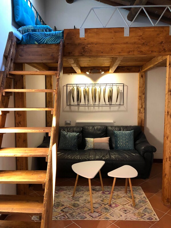 Ein Loft-Zimmer mit einer Holzleiter, einer schwarzen Ledersofa, zwei weißen Tischen und einem gemusterten Teppich.