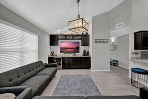Un salon moderne avec un canapé gris, un tapis bleu et un coin cuisine doté de meubles sombres et d’un grand écran de télévision diffusant une scène de coucher de soleil.