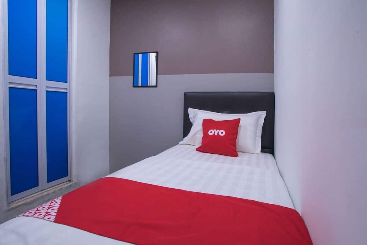 Une chambre d’hôtel avec un lit, une couvre-lit rouge et blanche, un oreiller rouge orné du logo « OYO », une fenêtre avec des rideaux bleus et un luminaire fixé au mur.