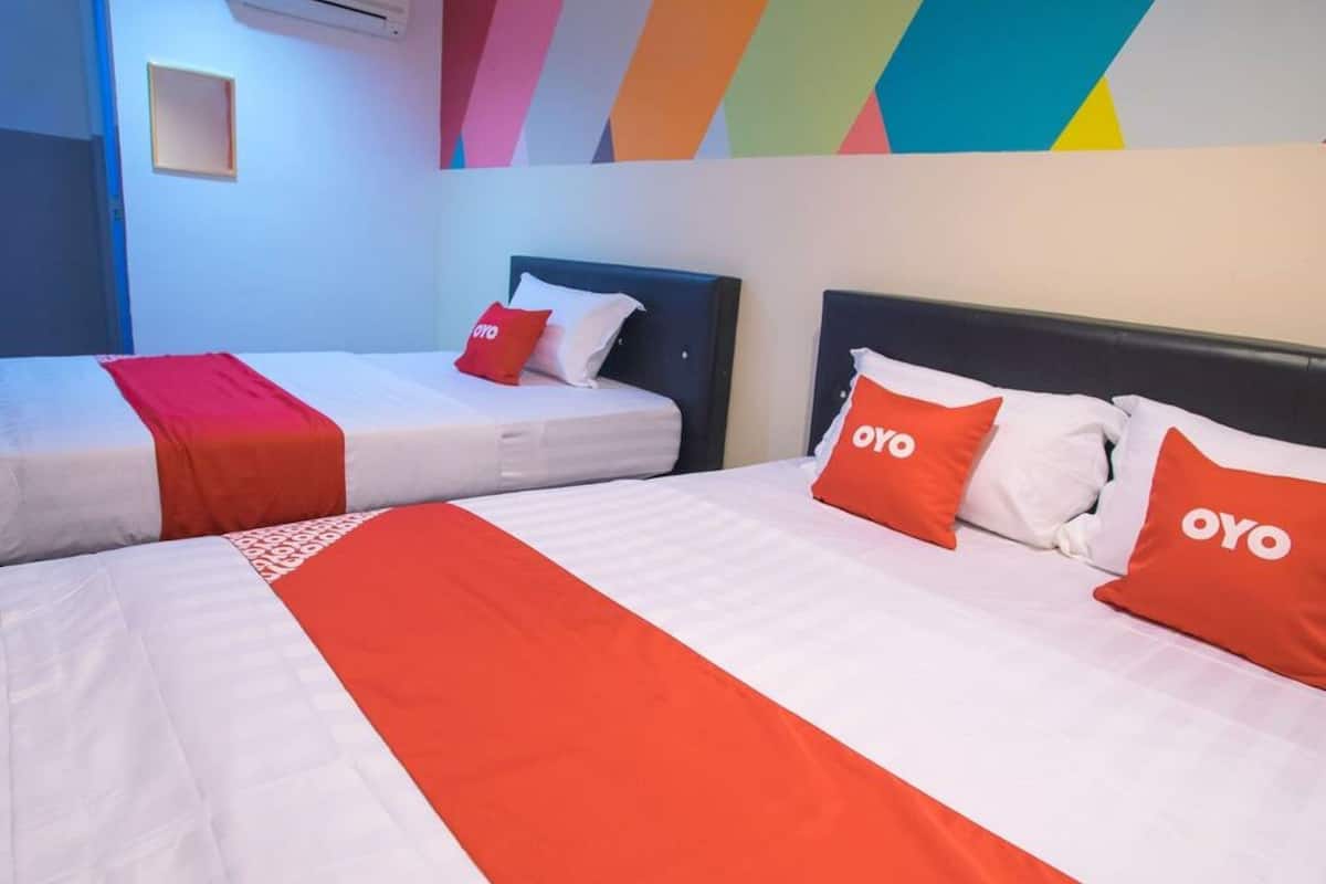 Une chambre d’hôtel avec deux lits, chacun agrémenté de draps blancs et de touches de couleur rouge, notamment des oreillers portant le nom de la marque « OYO ».
