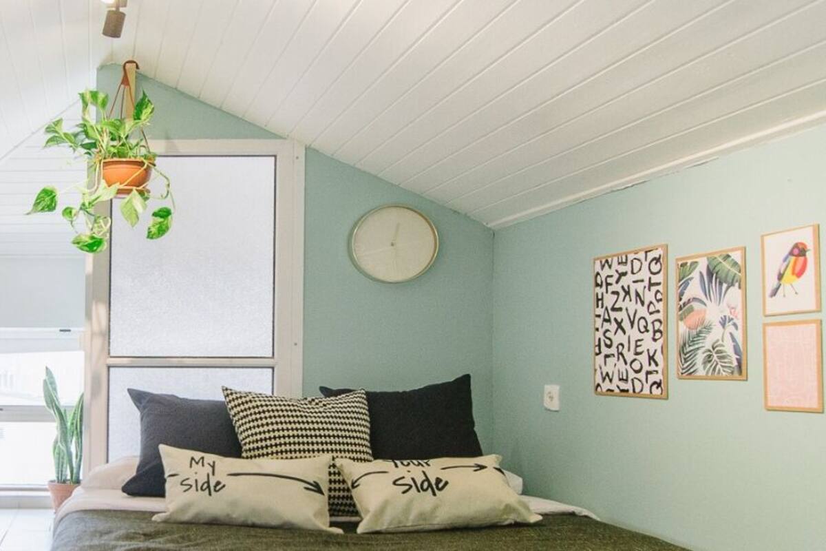 A cozy bedroom with a bed, pillows, a hanging plant, and a view of the kitchen.