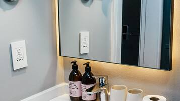 Un baño moderno con un lavamanos blanco, dos botellas de champú, dos tazas blancas y un rollo de papel higiénico.