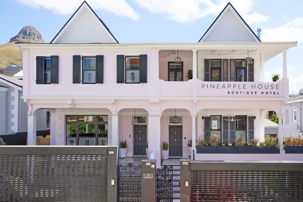 Un hôtel-boutique à deux étages, avec une façade rose, de nombreuses fenêtres dotées de volets et une enseigne indiquant « Pineapple House Boutique Hotel ».