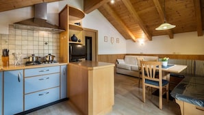 Cocina compacta con estufa a gas, armarios azules y vigas de madera. Se aprecia un comedor con mesa y sillas, y un sofá en el fondo.