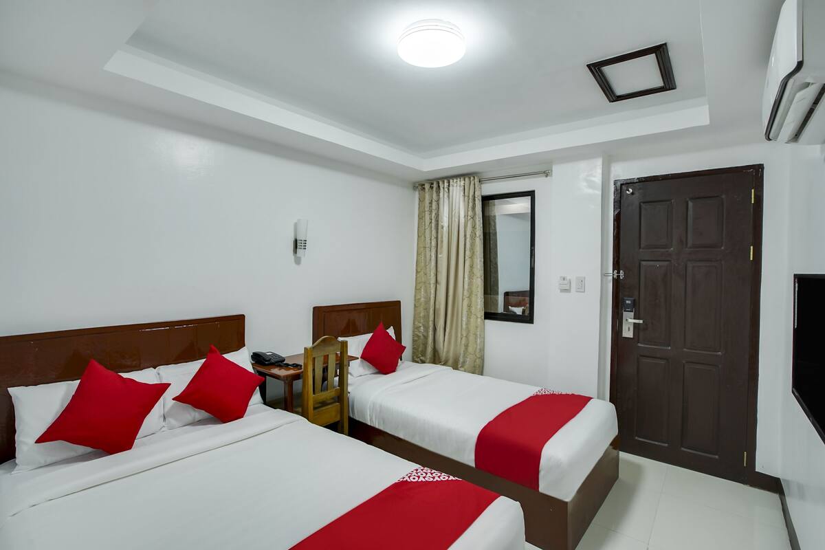 Quarto de hotel com duas camas, uma cabeceira de madeira, uma mesa com cadeira, telefone, janela com cortinas e uma porta.