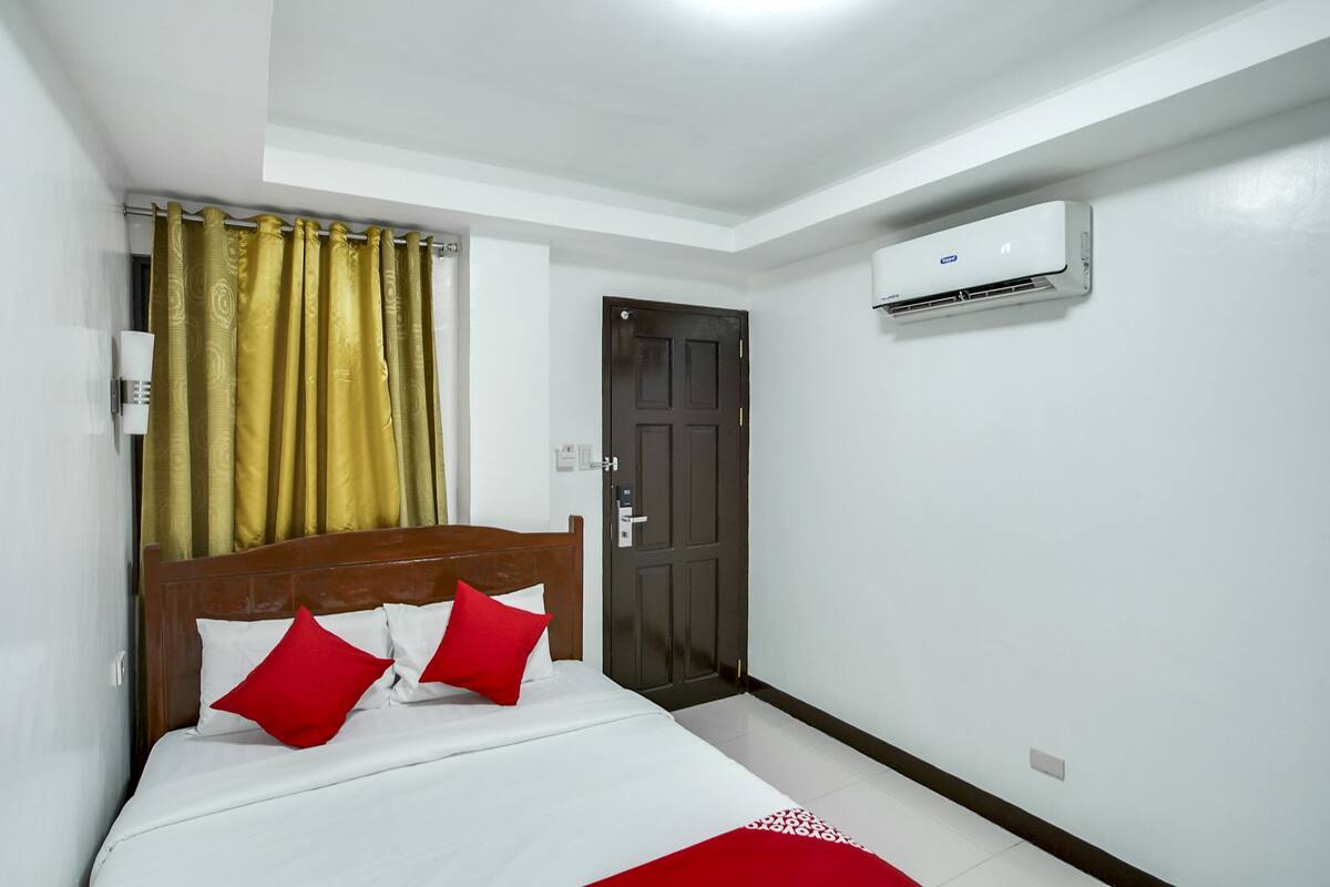 Quarto de hotel com cama, cortinas, porta e aparelho de ar condicionado.